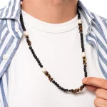 Thales è una collana da uomo realizzata a mano in Italia con pietre naturali di qualità, che donano un tocco di stile e personalità al tuo look, un accessorio che incanta per la sua moderna raffinatezza. Intrecciata con pietra lavica dall’aspetto poroso che contrasta con l’aulite bianca, questa creazione esclusiva, grazie anche all’occhio di tigre, evoca un senso di avventura e mistero.<br /> Queste pietre hanno anche dei significati simbolici: la pietra lavica rappresenta la forza e la trasformazione, mentre l’occhio di tigre stimola la fiducia e la protezione.<br /> Questa collana si abbina con naturalezza a outfit casual e sportivi e anche con colori caldi e neutri. È perfetta per le vacanze estive o per regalare un tocco casual al tuo look quotidiano in tutte le stagioni. ndossa Thales e lasciati trasportare in un viaggio di stile e libertà, dove ogni dettaglio racconta una storia senza tempo. <ul class="testo-prodotti"> <li class="spaziatura"><strong>Materiali</strong>: Minuterie e chiusura placcate argento 5 micron, ciuffo di bandana in cotone</li> <li class="spaziatura"><strong>Pietre</strong>: Pietra lavica con taglio a perla, aulite bianca a sasso e chips di occhio di tigre marrone</li> <li class="spaziatura"><strong>Taglia Unica</strong>: La lunghezza totale della collana è di 70cm</li> <li class="spaziatura"><strong>Chiusura</strong>: Con moschettone</li> <li class="spaziatura"><strong>Confezione</strong>: La collana è venduta all’interno di un’elegante scatolina con logo Dandy Street. Perfetta anche come regalo!</li> </ul>